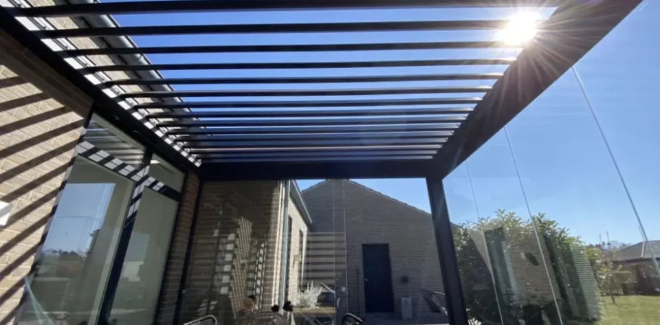 Grey frame bioclimatic pergola with glass sliding doors, furnished with a brown wooden dining table and black dining chairs.