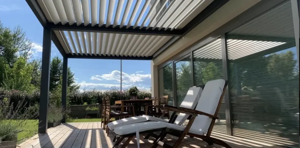 Grey frame bioclimatic pergola with white aluminium roof slats, furnished with white and brown sun loungers and brown wooden dining set.
