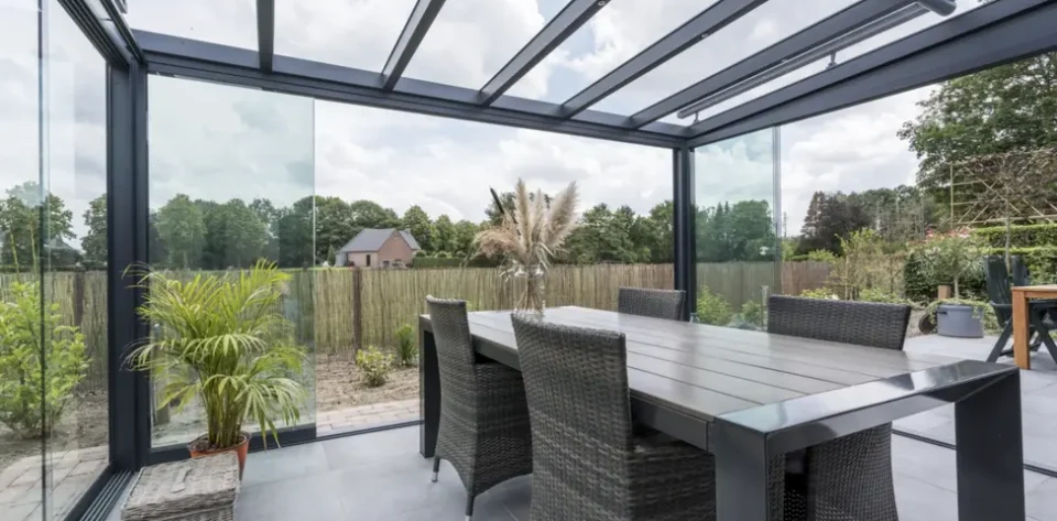 Anthracite grey frame glass room veranda with glass sliding doors and LED spot lights, furnished with dark beige rattan dining chairs and a dark brown dining table.