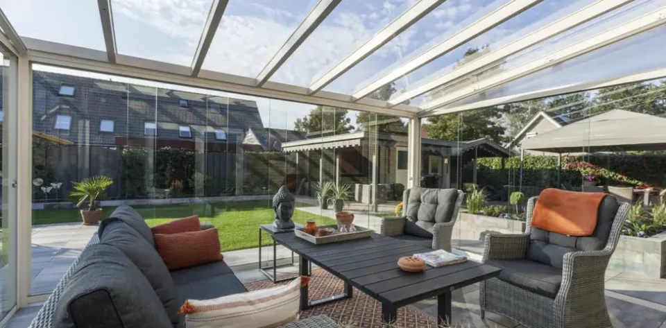 Silver bronze frame glass room veranda with glass sliding doors, furnished with beige rattan garden chair and sofa, brown wooden coffee table and orange pillows.